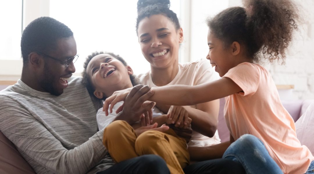 Overjoyed african American young family with little kids have fun playing relaxing on sofa in living room, happy biracial parents entertain with small children tickle laugh, enjoy weekend together