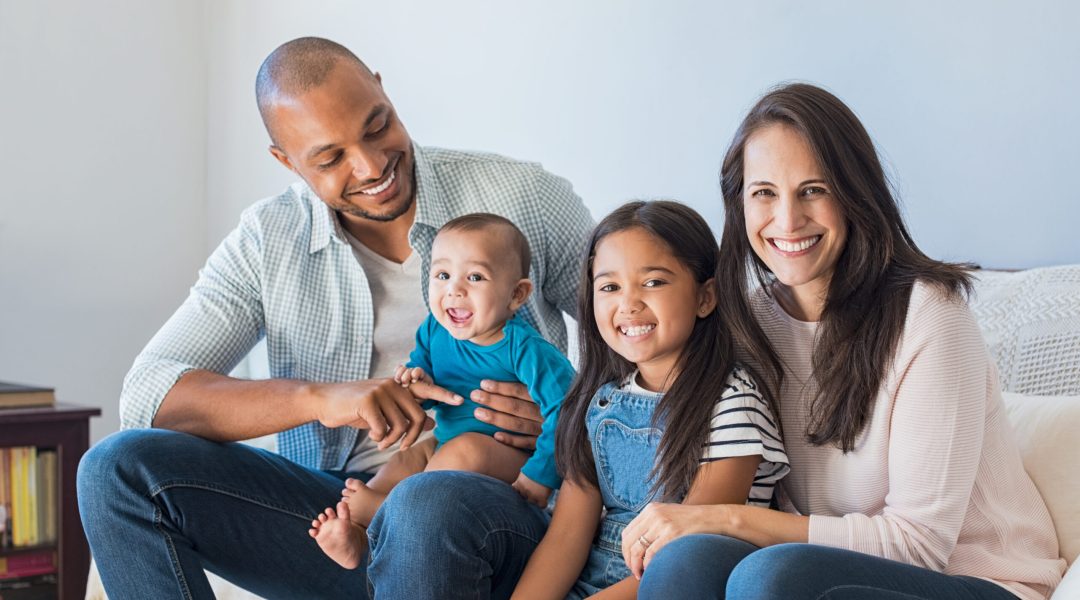 Portrait of happy multiethnic family sitting on sofa at home. Smiling couple with kids sitting on couch and looking at camera. Black father and latin woman with daughter sitting on couch and having fun.